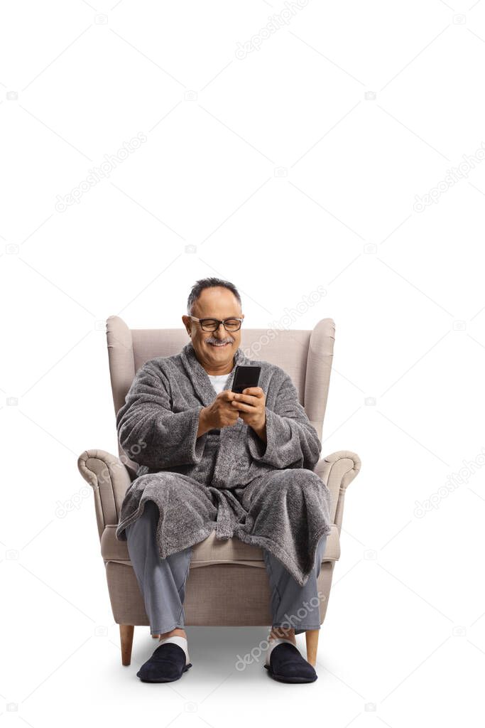 SMiling mature man in a bathrobe sitting in an armchair and typing on a smartphone isolated on white background