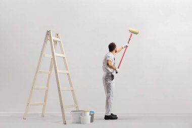 Merdiven ve kovalar bir odada ve dekoratör beyaz arka planda izole bir duvar boyuyor.