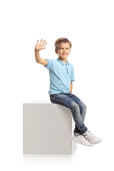 笑顔の男の子は白い立方体に座って 白い背景に隔離されたカメラで手を振る — ストック写真