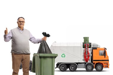 Olgun bir adam çöp kutusuna plastik poşet atıyor ve beyaz arka planda izole edilmiş bir çöp kamyonunun önünde baş parmağını gösteriyor.