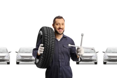 Beyaz arka planda izole edilmiş gümüş arabaların önünde bijon anahtarı ve lastik tutan oto tamircisi.