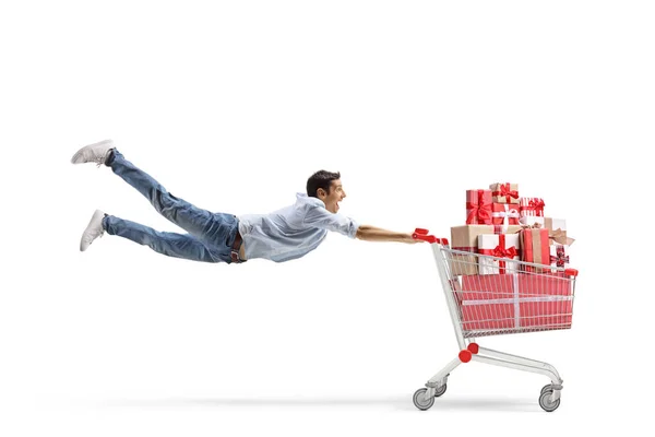 白い背景に隔離されたプレゼントでいっぱいのショッピングカートで飛ぶカジュアルな若い男の完全な長さのショット — ストック写真