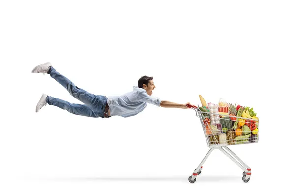 白い背景に孤立した食べ物とショッピングカートで飛ぶカジュアルな若い男の完全な長さのショット — ストック写真