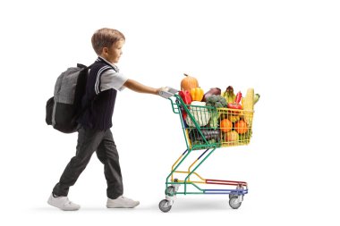 Okul üniformalı bir çocuğun, arka planda izole edilmiş meyve ve sebzelerle dolu mini bir alışveriş arabasını iterken tam boy profil fotoğrafı.