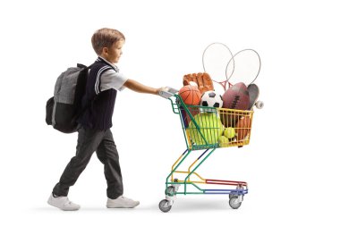 Tam boy profil fotoğrafı, beyaz bir grupta izole edilmiş spor ekipmanlarıyla küçük bir alışveriş arabasını iten bir okul çocuğu.