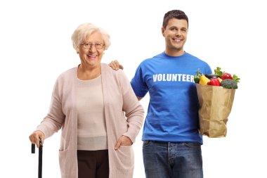 Yaşlı bir kadın, beyaz arka planda alışveriş çantasını taşıyan genç bir gönüllü ile ayakta duruyor.