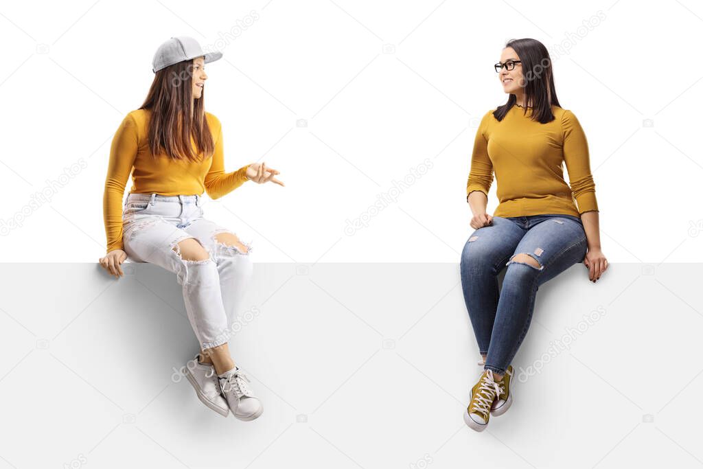Two young female friends sitting on a blank panel and having a conversation isolated on white background