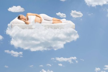 Pijamalı hamile bir kadın yatakta uyuyor ve gökyüzünde bulutların üzerinde süzülüyor.