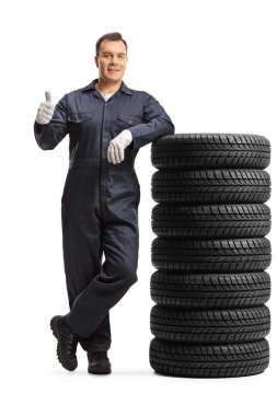 Üniformalı bir otomobil tamircisi lastik yığınına yaslanıyor ve beyaz arka planda izole edilmiş bir baş parmak hareketi gösteriyor.
