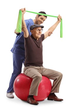 Physical therapist working with an elderly man sitting on an exercise ball and using a stretching band isolated on white background