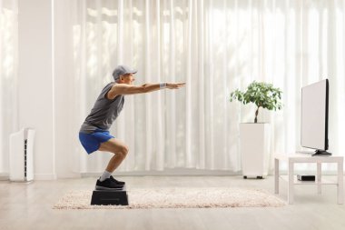 Full length profile shot of an elderly man exercising squats on a step aerobic platform in front of tv