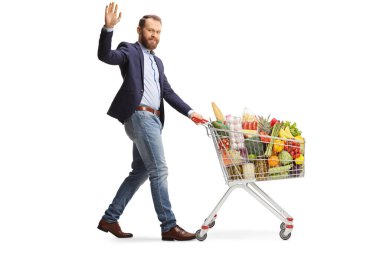 Takım elbiseli sakallı genç bir adam alışveriş arabasıyla yürüyor ve beyaz arka planda izole bir şekilde el sallıyor.