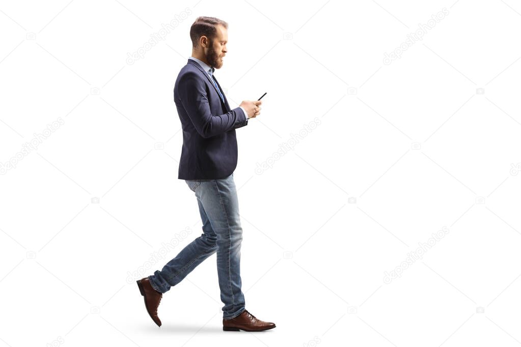 Full length profile shot of a young man in suit and jeans walking and typing on a mobile phone isolated on white background