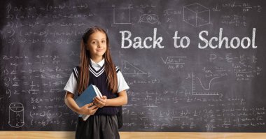 Okul üniformalı bir öğrenci, elinde karatahtanın önünde bir kitap tutuyor ve okula geri dönüyor.