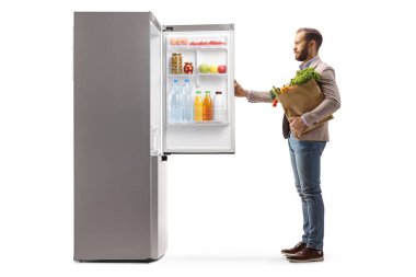 Bir adamın buzdolabını açarken ve elinde beyaz arka planda izole edilmiş bir poşet tutarken tam boy profil fotoğrafı.