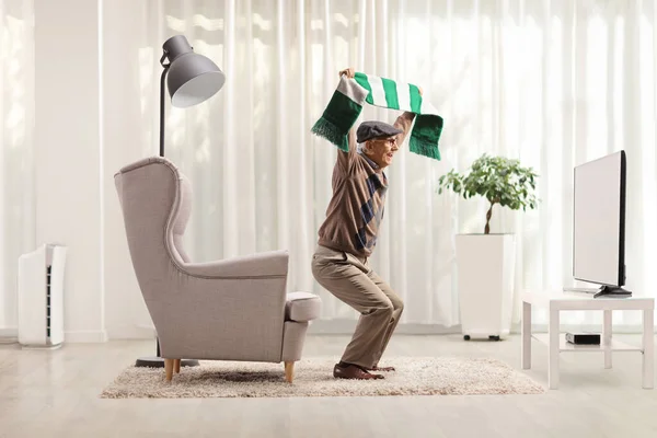 拿着足球围巾的老人在家里一边欢呼一边看电视上的比赛 — 图库照片
