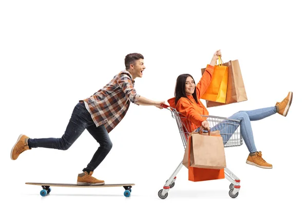 男オンAスケートボードプッシュ若い女性でショッピングカートの中でショッピングバッグで隔離された白い背景 — ストック写真