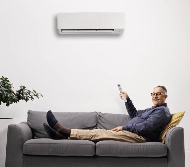 Olgun bir adam kanepeye uzanıyor ve beyaz arka planda izole edilmiş uzaktan kumandayla klimayı açıyor.