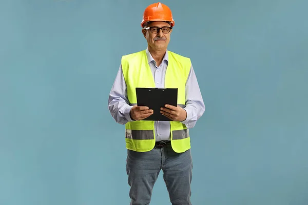 男性成熟工程师 身穿安全背心 头戴安全帽 手持蓝色背景隔离的剪贴板 — 图库照片