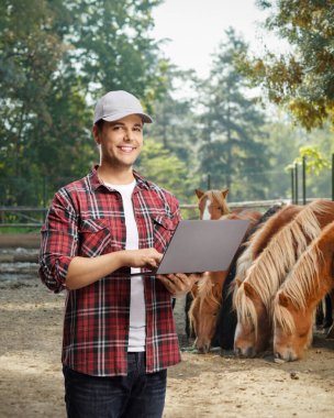 Laptop bilgisayarı olan çiftçi gülümseyen ve poz veren bir aygır çiftliğinde