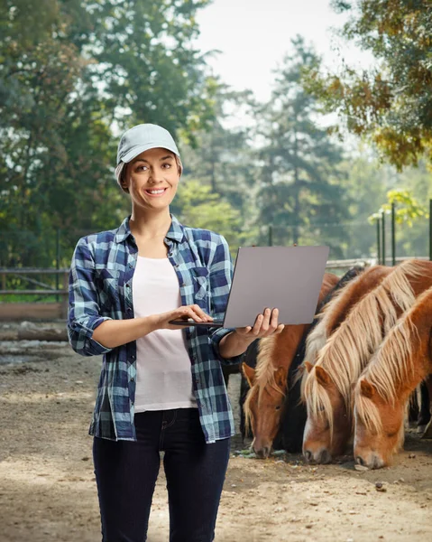 背中に馬とスタッドの農場に笑顔とポーズラップトップコンピュータを持つ現代の女性農家 — ストック写真