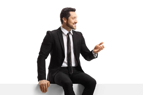 Young man in a suit sitting on a blank panel and gesturing with hand isolated on white background