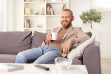 Genç adam gülümsüyor ve evinde bir fincan çayla kanepede oturuyor.