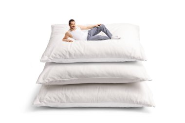 Beyaz arka planda izole edilmiş büyük yastıkların üzerinde pijamalı genç bir adam yatıyor.