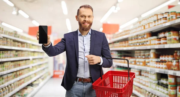 Smiling Young Man Holding Shopping Basket Showing Mobile Phone Supermarket — Stockfoto