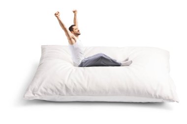 Pijamalı genç adam beyaz arka planda büyük bir yastığa uzanıyor ve oturuyor.