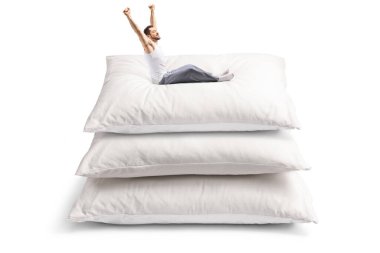 Pijamalı genç adam beyaz arka planda izole edilmiş büyük yastıkların üzerinde uzanıyor ve oturuyor.