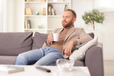 Young man sitting on a sofa at home with a cup of tea and thinking