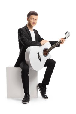 Akustik gitarlı siyah takım elbiseli genç adam beyaz arka planda izole edilmiş beyaz bir küpün üzerinde oturuyor.