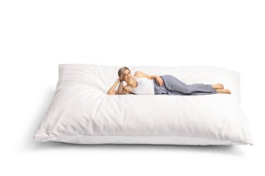 Güzel genç bir kadın, rahat bir yastığın üzerinde uzanıyor. Beyaz arka planda izole edilmiş pijamalarıyla.