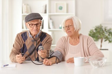 Yaşlı koca karısına kan basıncını ölçüyor ve evde bir masada oturuyor.