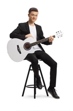 Siyah takım elbiseli zarif genç bir adam bir sandalyede oturuyor ve beyaz arka planda izole edilmiş beyaz bir akustik gitar tutuyor.
