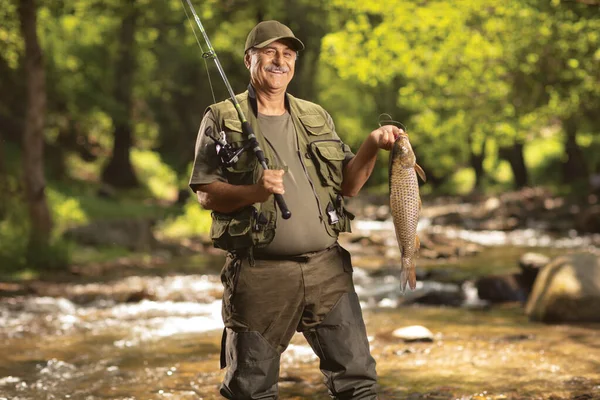 拿着鲤鱼 站在林子里的河里 微笑着成熟的渔夫 — 图库照片