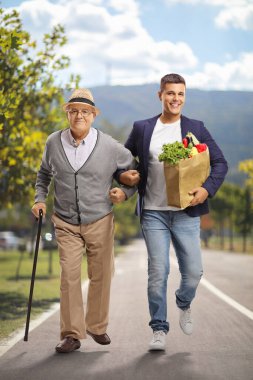 Genç bir adam yaşlı bir adama market çantasıyla yardım ediyor ve yaya yolunda yürüyor.