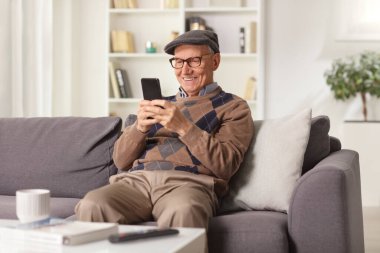 Yaşlı adam gülümsüyor ve akıllı telefon kullanıyor. Evdeki kanepede oturuyor. 