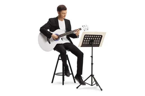 身穿黑色西服的年轻人坐在椅子上 弹着一把带着乐谱的音响吉他 背景是白色的 — 图库照片
