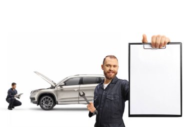 Tamirci, elinde İngiliz anahtarı ve beyaz arka planda izole edilmiş boş bir kağıt tutan üniformalı bir aracı ve diğer işçiyi kontrol ediyor.