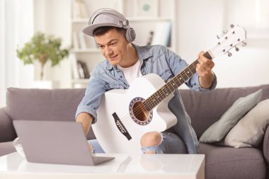 Evdeki koltukta oturan, akustik gitar çalan ve dizüstü bilgisayara bakan bir adam.