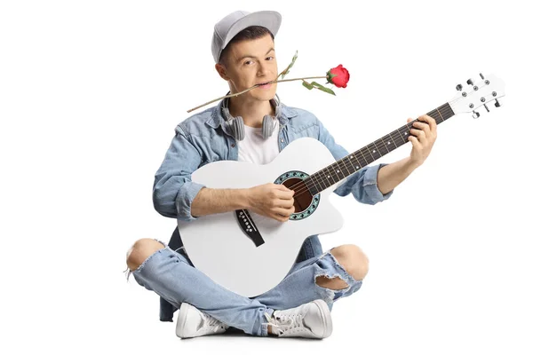 年轻男子 嘴里衔着玫瑰 坐在地板上弹奏着声吉他 背景是白色的 — 图库照片