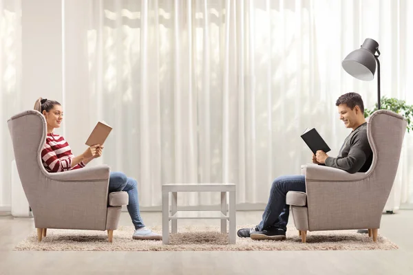 プロフィールショットの若い男と女に座っていますアームチェアと読書本に家 — ストック写真