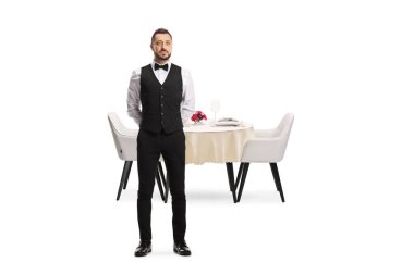 Beyaz arka planda izole bir masanın önünde duran papyonlu bir garsonun tam boy portresi.
