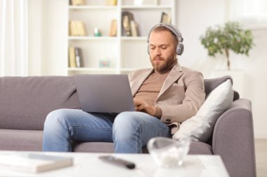 Kulaklıklı bir adam dizüstü bilgisayar kullanıyor ve evdeki kanepede oturuyor. 
