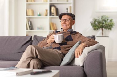 Yaşlı bir adam evdeki kanepede dinleniyor ve elinde bir fincan çay tutuyor.
