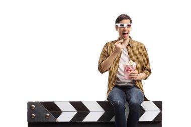 Üç boyutlu gözlüklü bir adam film sehpasında oturup beyaz arka planda izole edilmiş patlamış mısır yiyor.