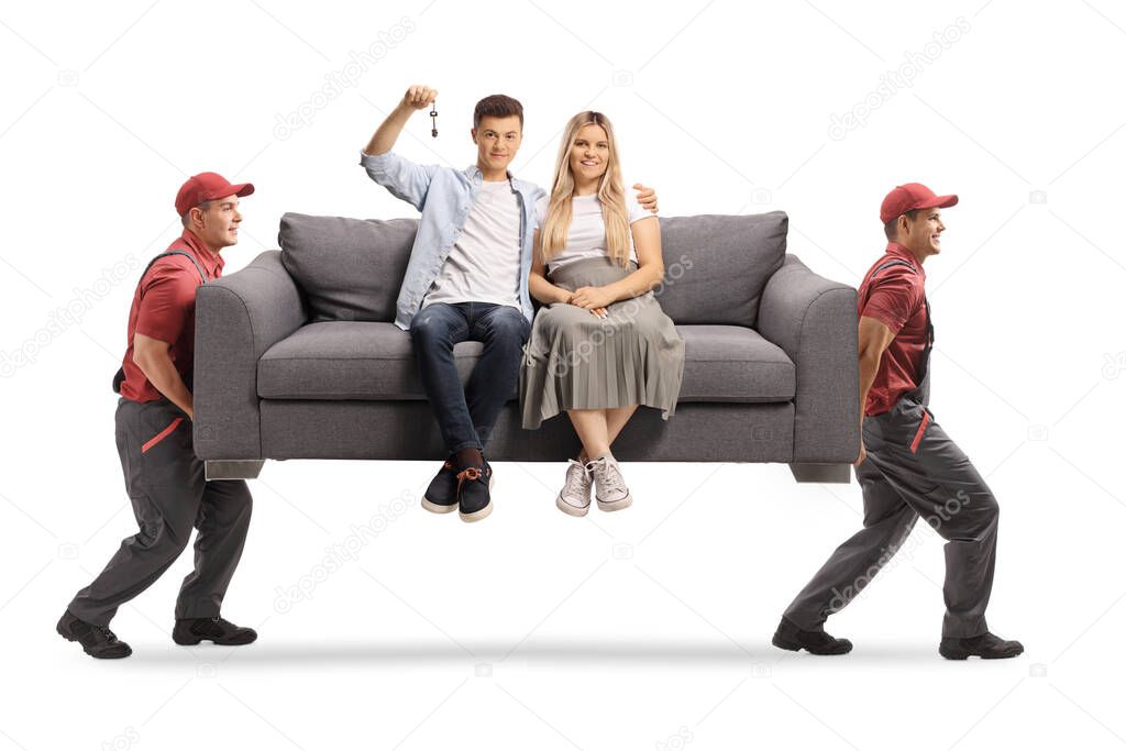 Movers carrying a young couple sitting on a gray sofa and holding a house key isolated on white background
