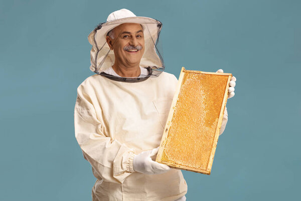Mature male bee keeper in a uniform holding a honeybee frame isolated on blue background